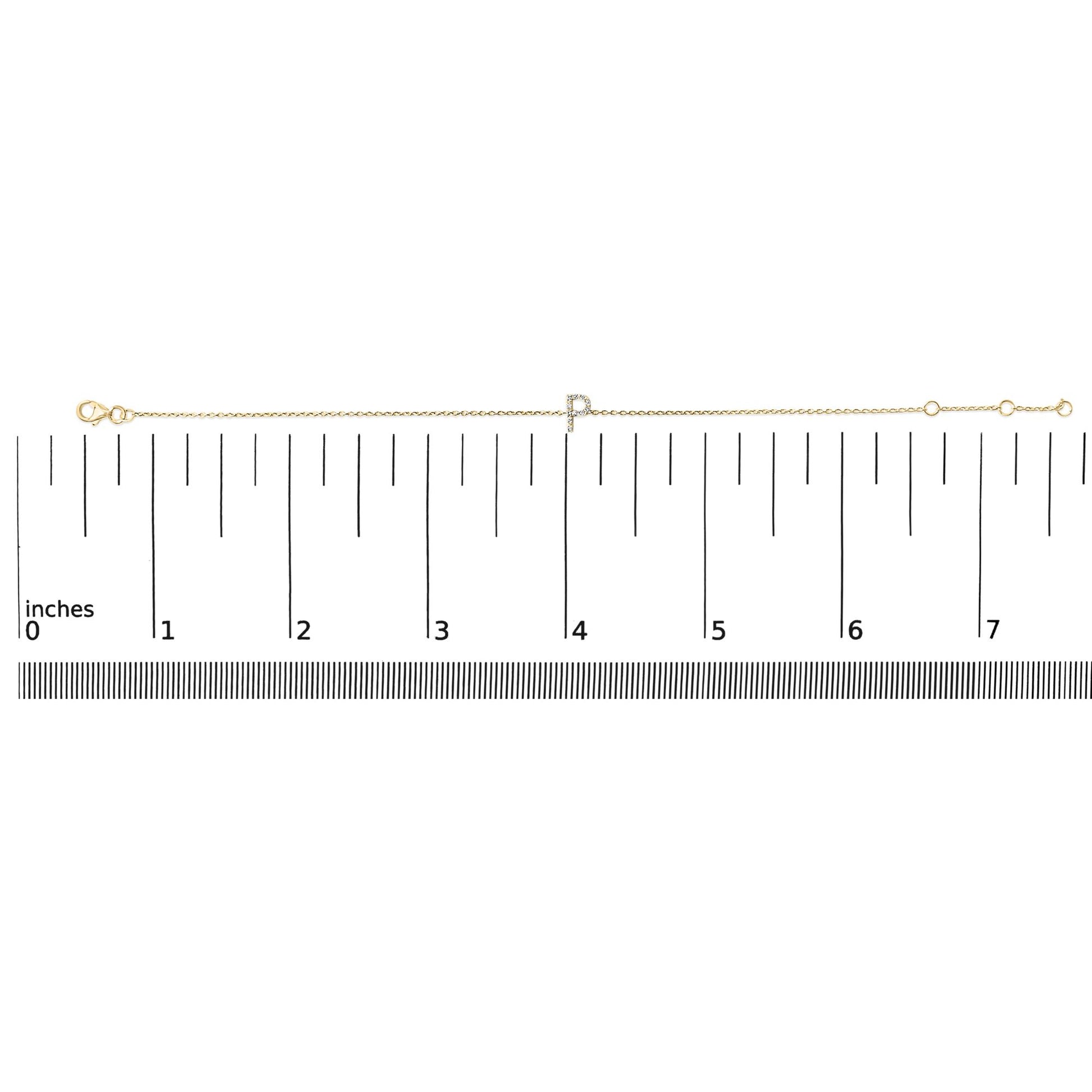 Yellow metal bracelet with a micro-pave set white gemstone initial charm, shown on a ruler scale from a top-down angle.