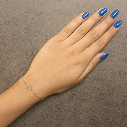 Yellow bracelet with a delicate chain and a small letter charm set with white round gemstones, shown worn on a model's wrist, top-down angle.