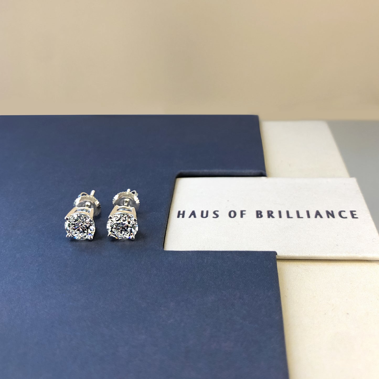 Close-up front angle of a pair of white metal stud earrings with round white gemstones in four-prong settings, shown lying on a blue surface next to a card with the text 'HAUS OF BRILLIANCE.'