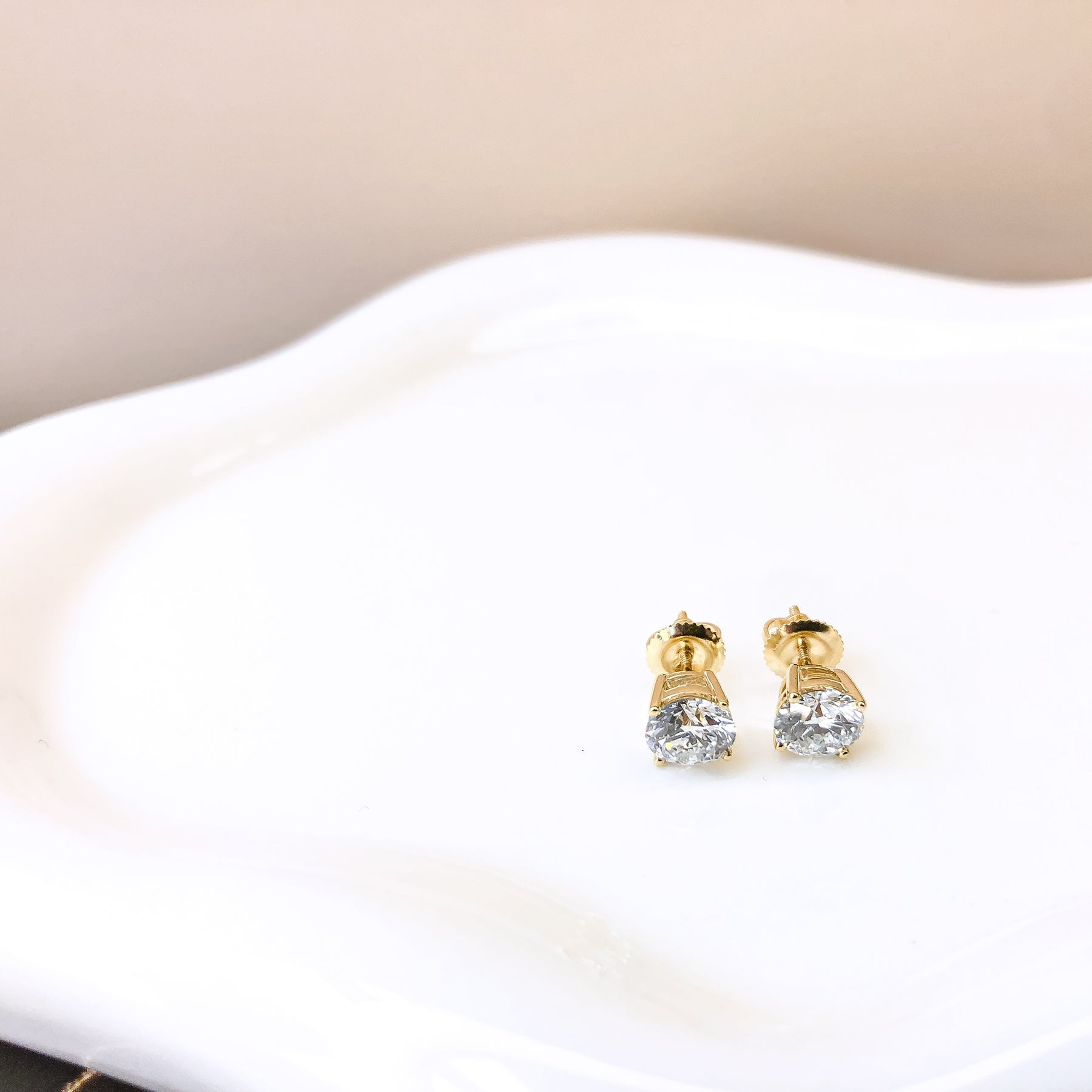 Pair of yellow metal stud earrings with round white gemstones in four-prong settings, shown from a front angle on a white dish.