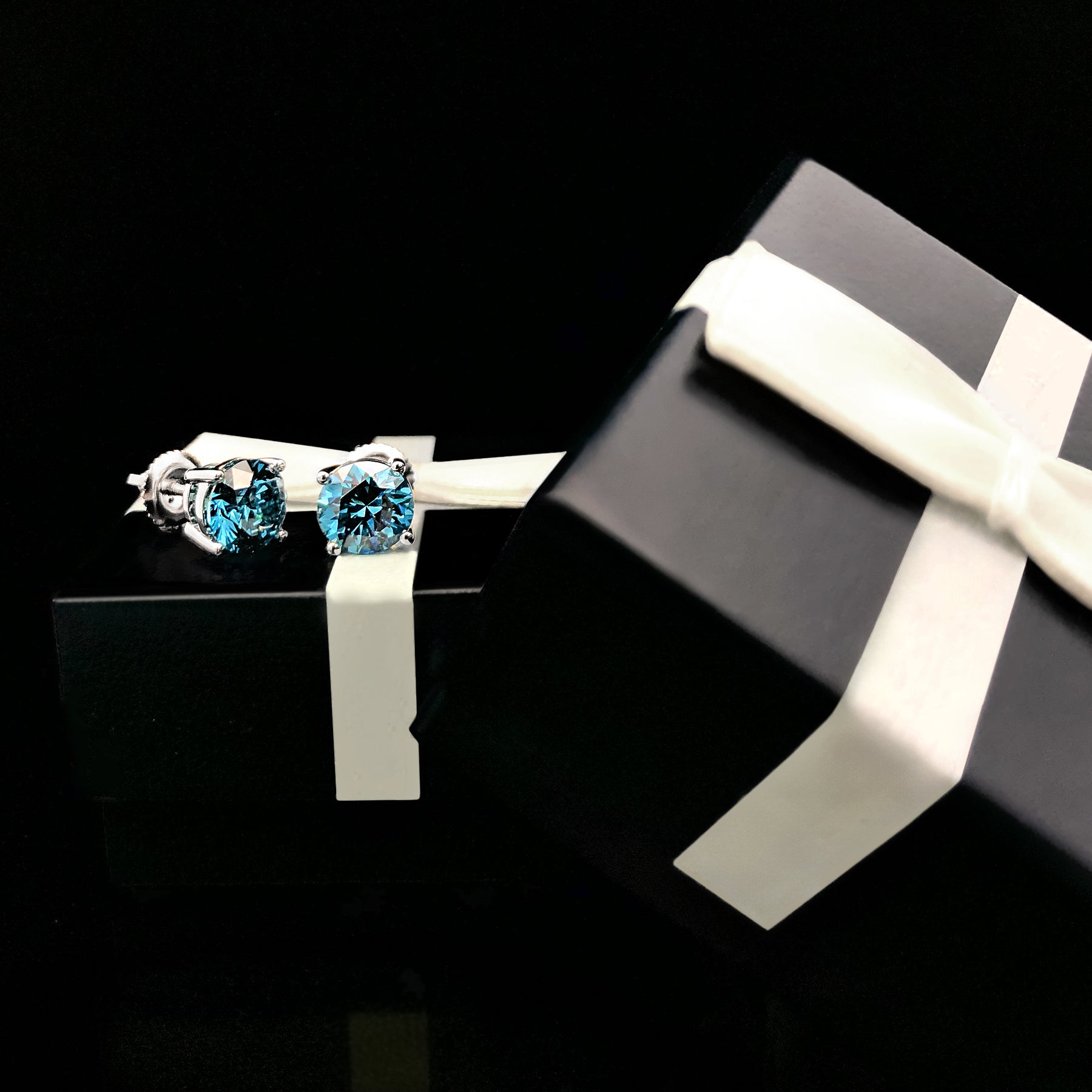 Pair of white metal round blue gemstone stud earrings in 4-prong settings, shown at an angle resting on a black gift box with a white ribbon.