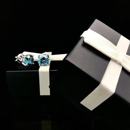 Pair of white metal round blue gemstone stud earrings in 4-prong settings, shown at an angle resting on a black gift box with a white ribbon.