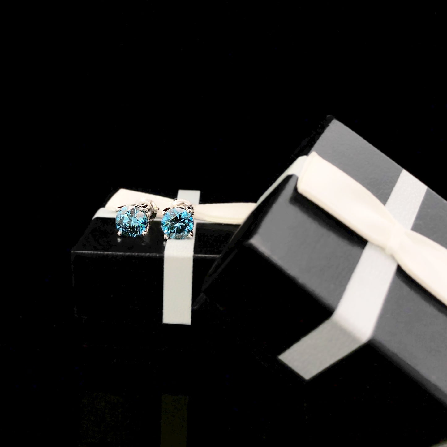 Pair of white metal stud earrings with round blue gemstones in 4-prong settings, shown on a black gift box with white ribbon, angled view.