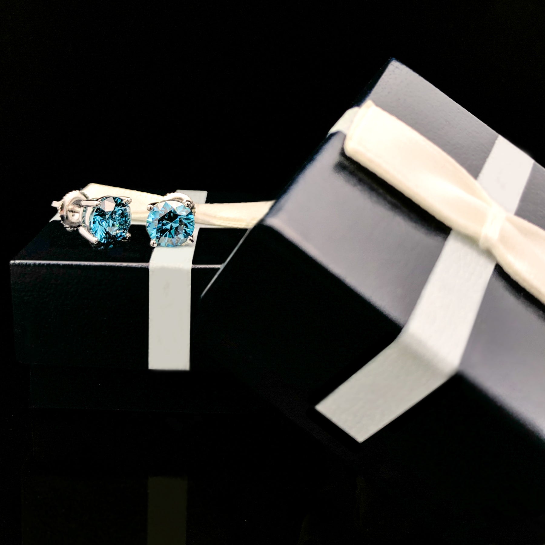 Close-up angled view of a pair of white metal stud earrings with round blue gemstones in four-prong settings, displayed on a black gift box with a white ribbon.