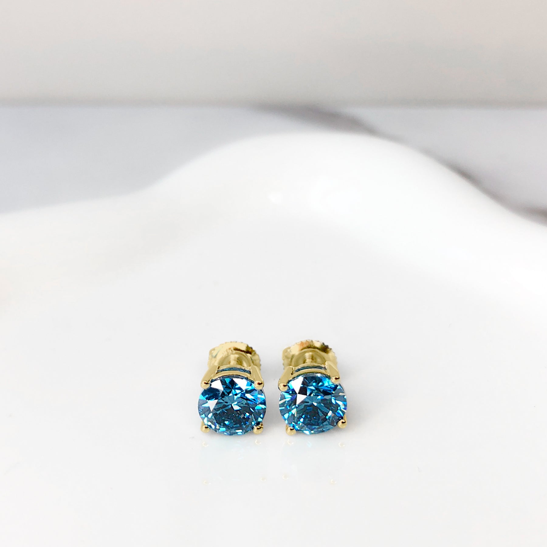 Pair of yellow metal stud earrings with round blue gemstones in four-prong settings, shown from the front angle on a white surface.