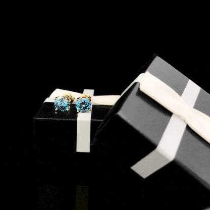 Close-up front angle of yellow metal stud earrings with round blue gemstones in 4-prong settings, displayed on a black gift box with a white ribbon.