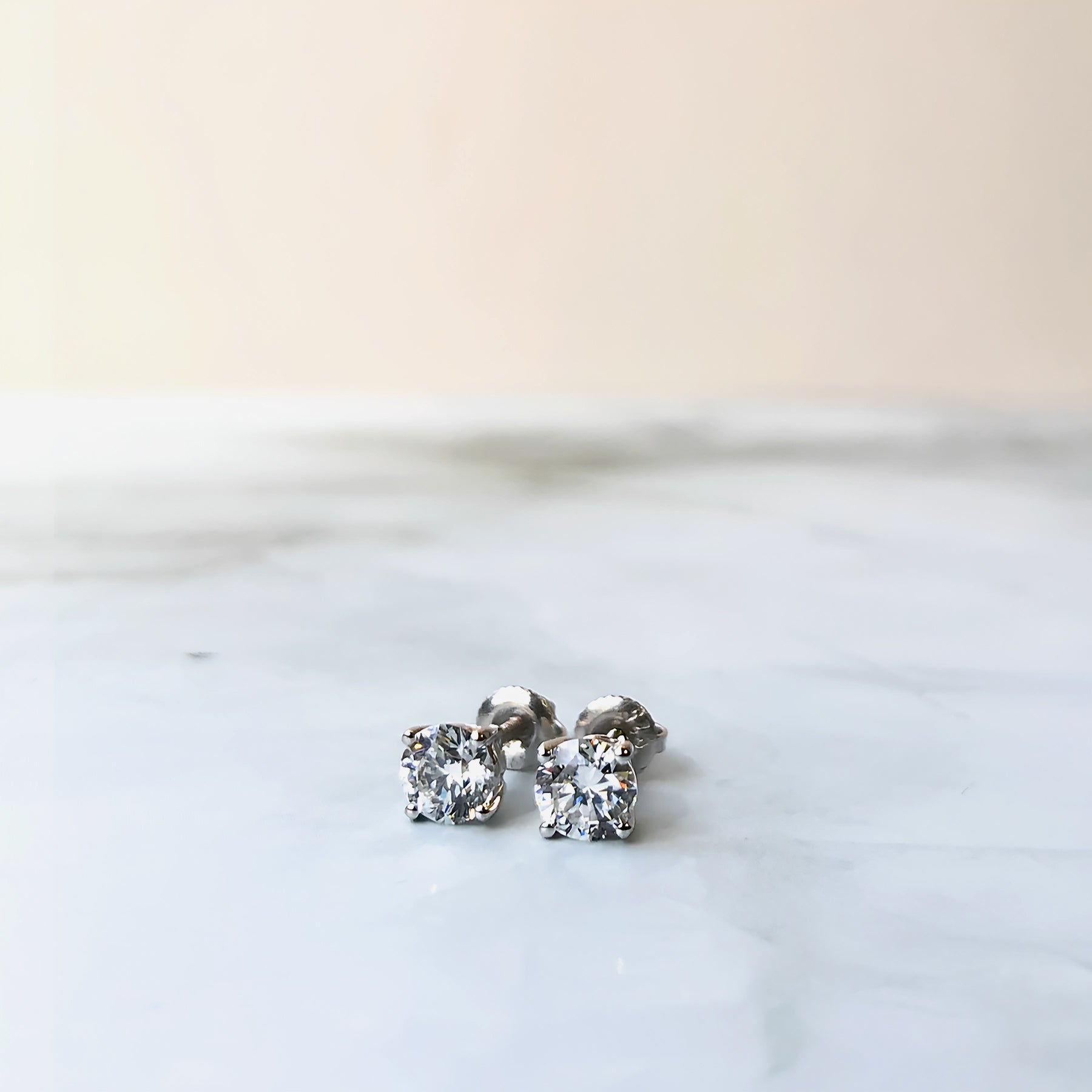 Close-up front angle of a pair of white metal stud earrings, each featuring a single round white gemstone in a four-prong setting, shown resting on a white marble surface.