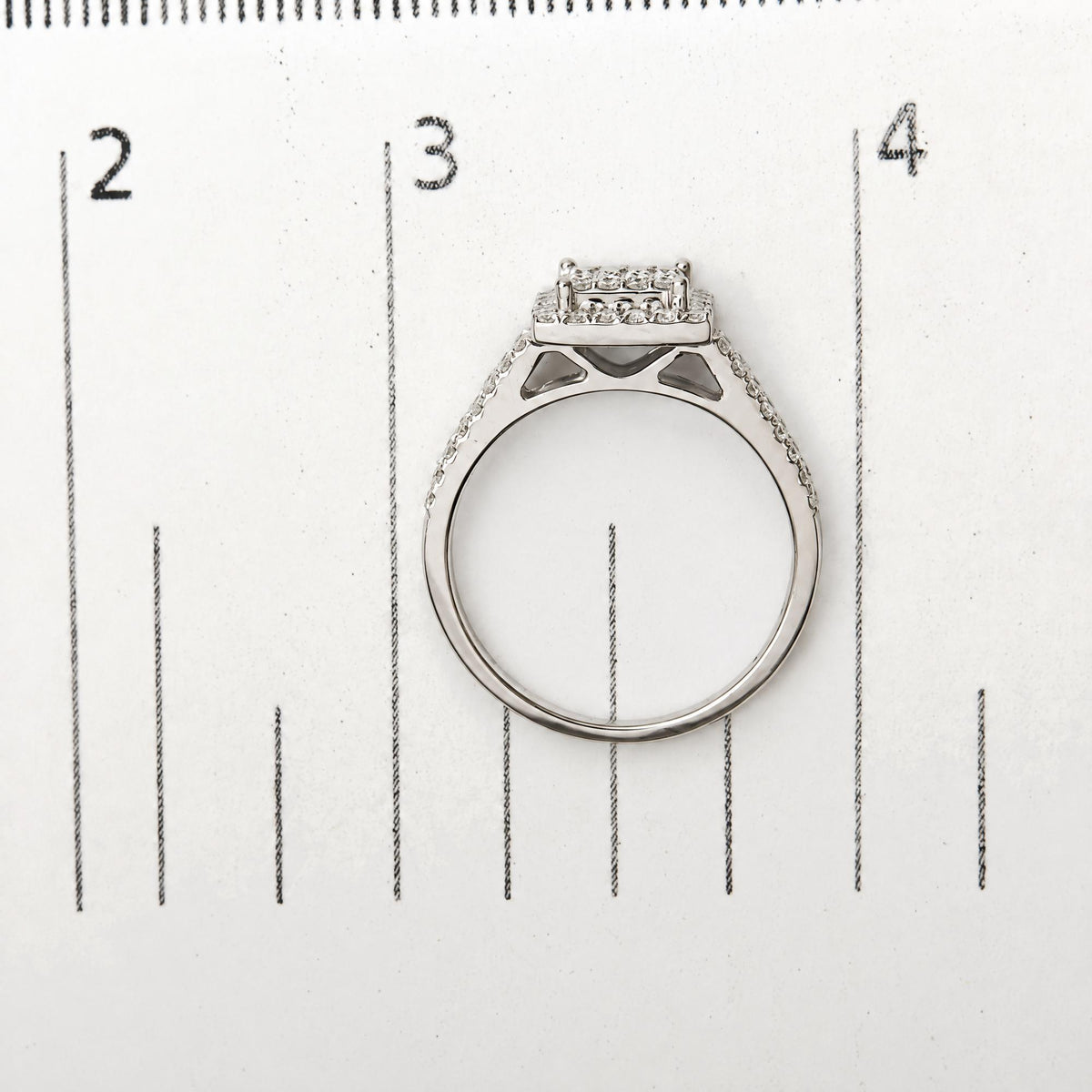 Top view of a white metal ring with round white gemstones in prong settings, shown on a measuring scale.