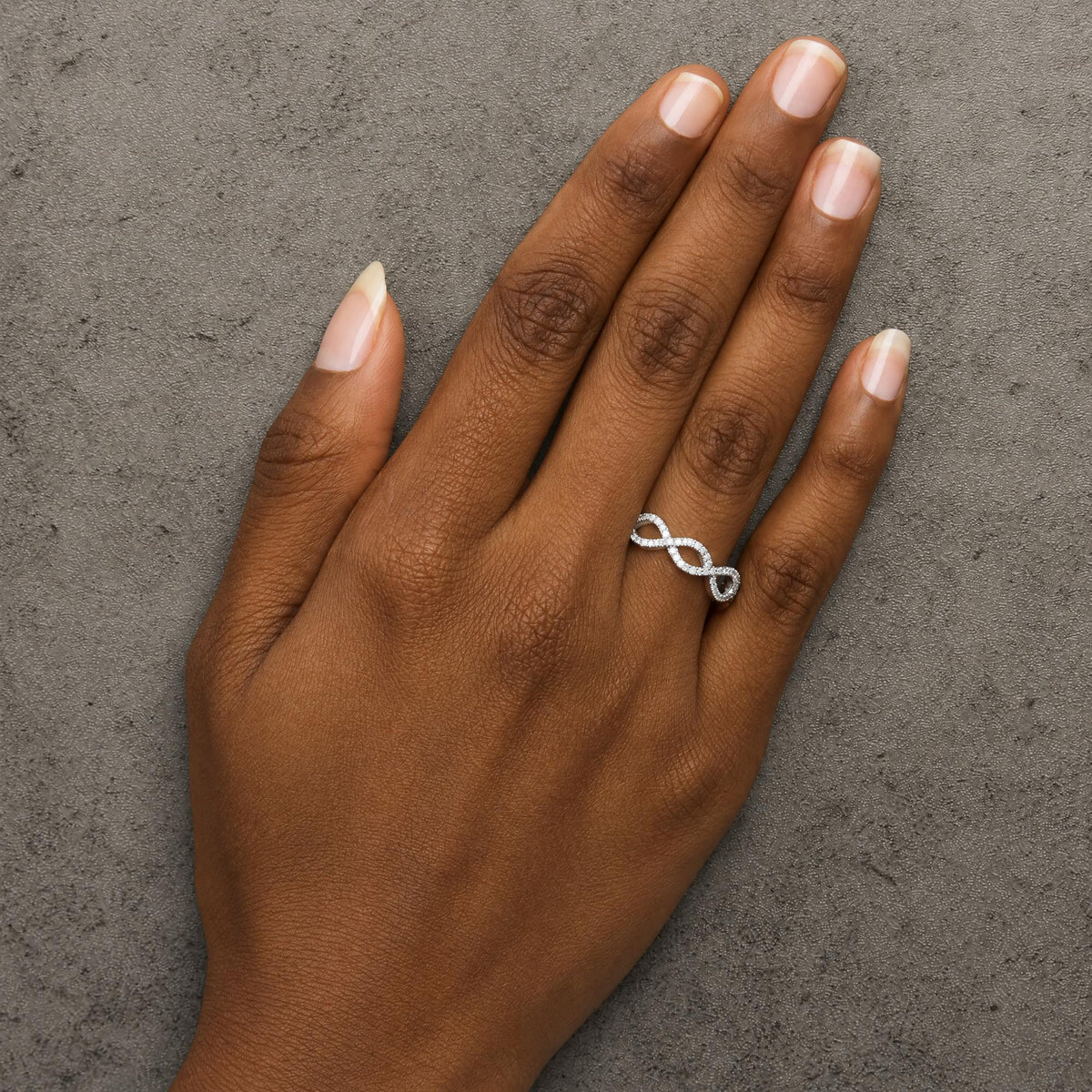 White metal infinity-shaped ring with round white gemstones in prong settings, shown worn on a model's hand from a top-down angle.