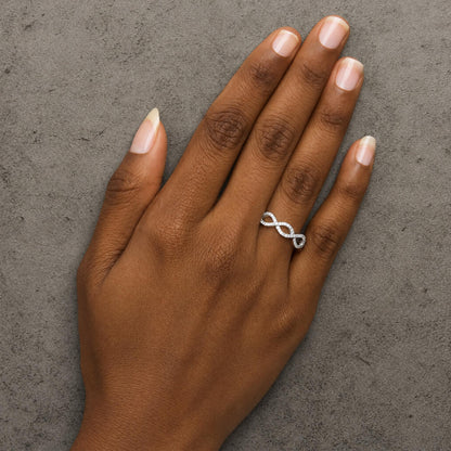 White metal infinity-shaped ring with round white gemstones in prong settings, shown worn on a model's hand from a top-down angle.