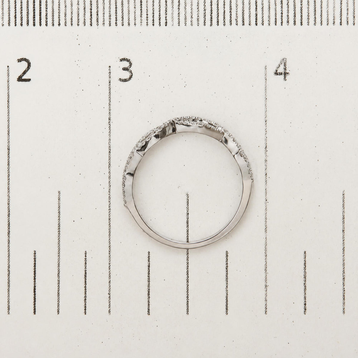 White ring with small round white gemstones in prong setting encircling the band, shown flat on a ruler for scale, viewed from above.