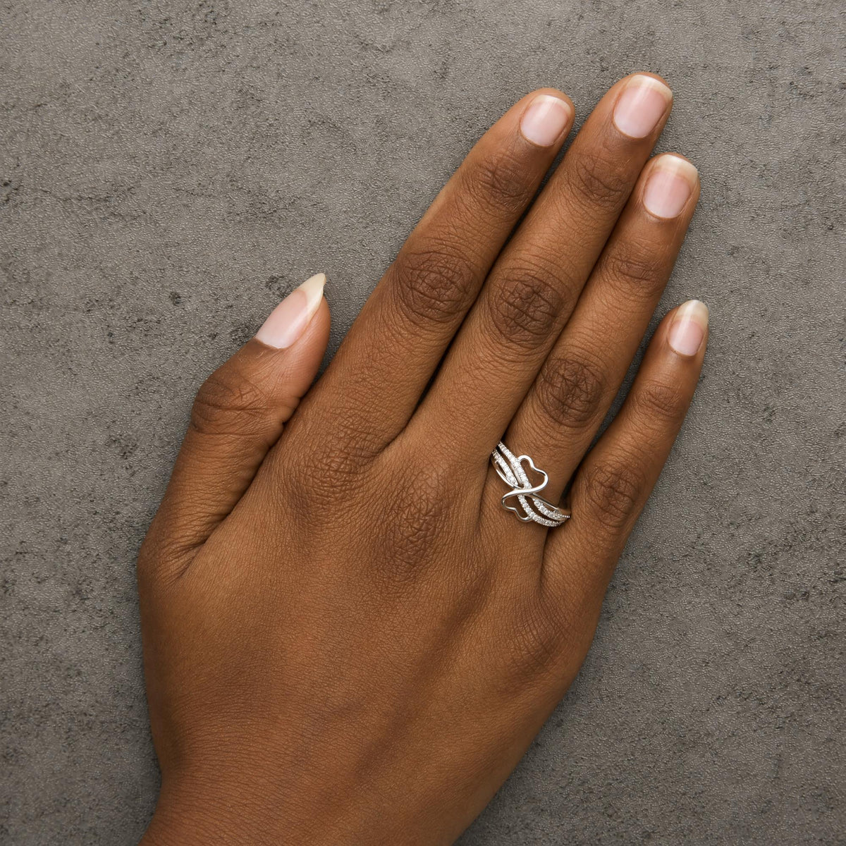 White metal ring with multiple intertwined bands adorned with round white gemstones in prong settings, shown worn on a model’s hand from a top-down angle.