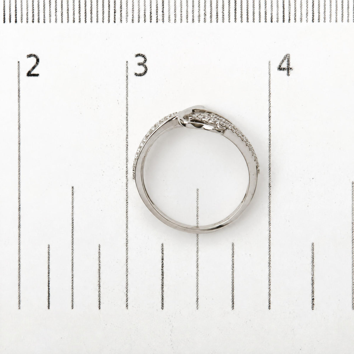 White ring with two rows of small round white gemstones in prong settings crossing at the top, shown on a ruler scale, top flat angle.