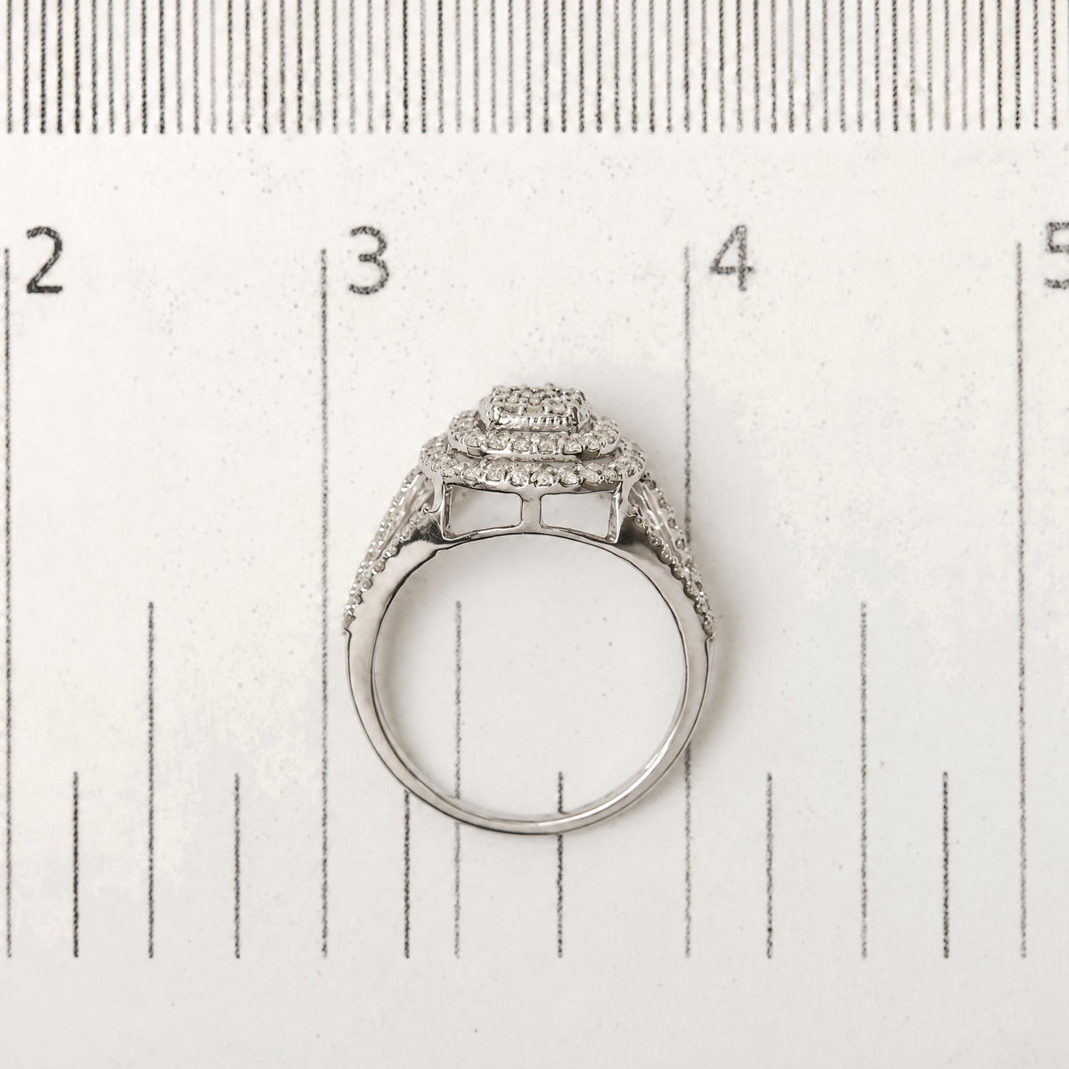 Close-up top view of a white metal ring with multiple rows of small round white gemstones in prong settings, shown on a scale.