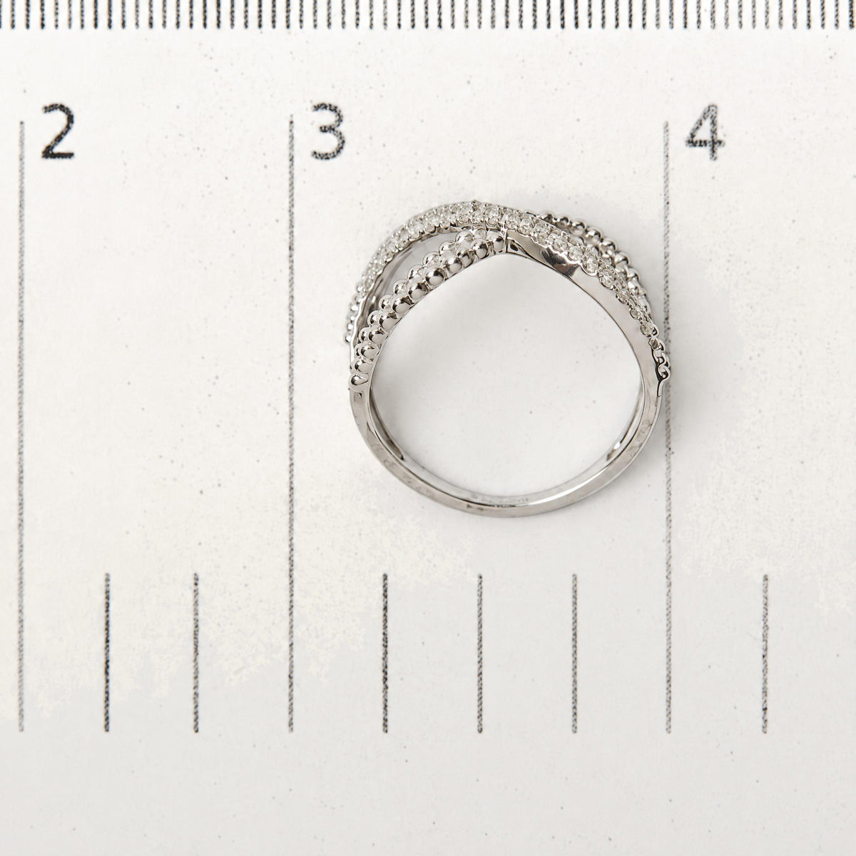 Close-up top view of a white metal ring with two overlapping bands, one featuring round white gemstones in prong settings and the other with smooth rounded beads, shown on a ruler for scale.