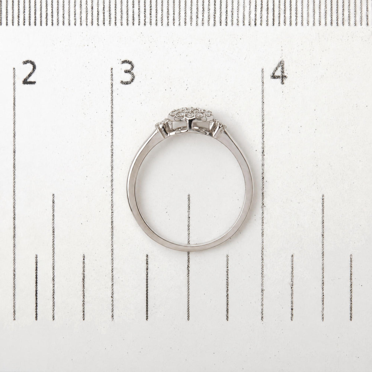 Close-up of a white metal ring with round white gemstones in prong settings arranged in a cluster, shown on a measurement scale, viewed from the side and slightly above.