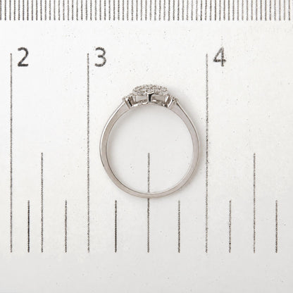 Close-up of a white metal ring with round white gemstones in prong settings arranged in a cluster, shown on a measurement scale, viewed from the side and slightly above.