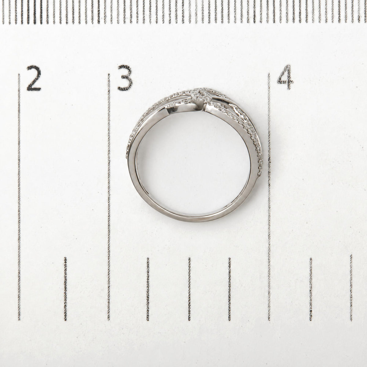 Close-up top view of a white metal ring with a split shank design set with round white gemstones in prong settings, placed next to a ruler for scale.