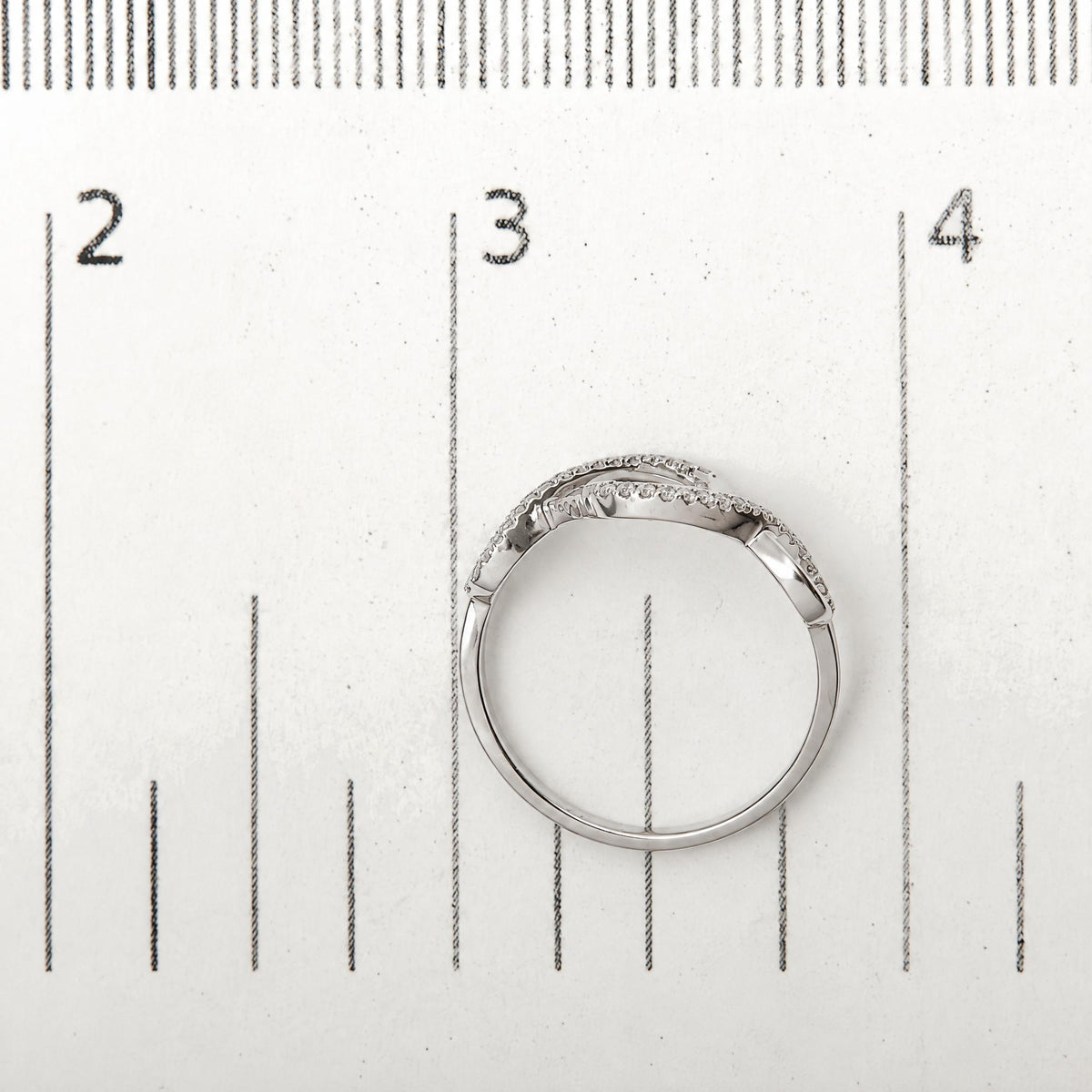 Close-up image of a white metal ring with two intertwined bands, each set with round white gemstones in prong settings, shown on a measurement scale.
