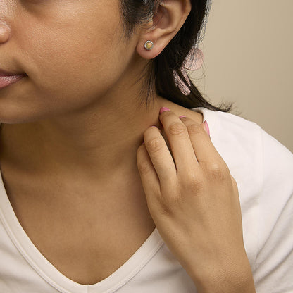 Close-up side view of a model wearing a yellow metal stud earring featuring a round multi-color opal gemstone in a four-prong setting, surrounded by a halo of small round white gemstones in a shared prong setting.