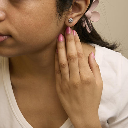 Close-up side view of a model wearing round white gemstone stud earrings surrounded by smaller white gemstones in a shared prong setting, with the model's hand touching her ear.