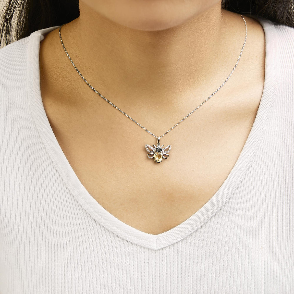 White metal necklace shown worn on a model, featuring a bee-shaped pendant with an oval orange gemstone, an oval black gemstone, and multiple round white gemstones, photographed from the front.