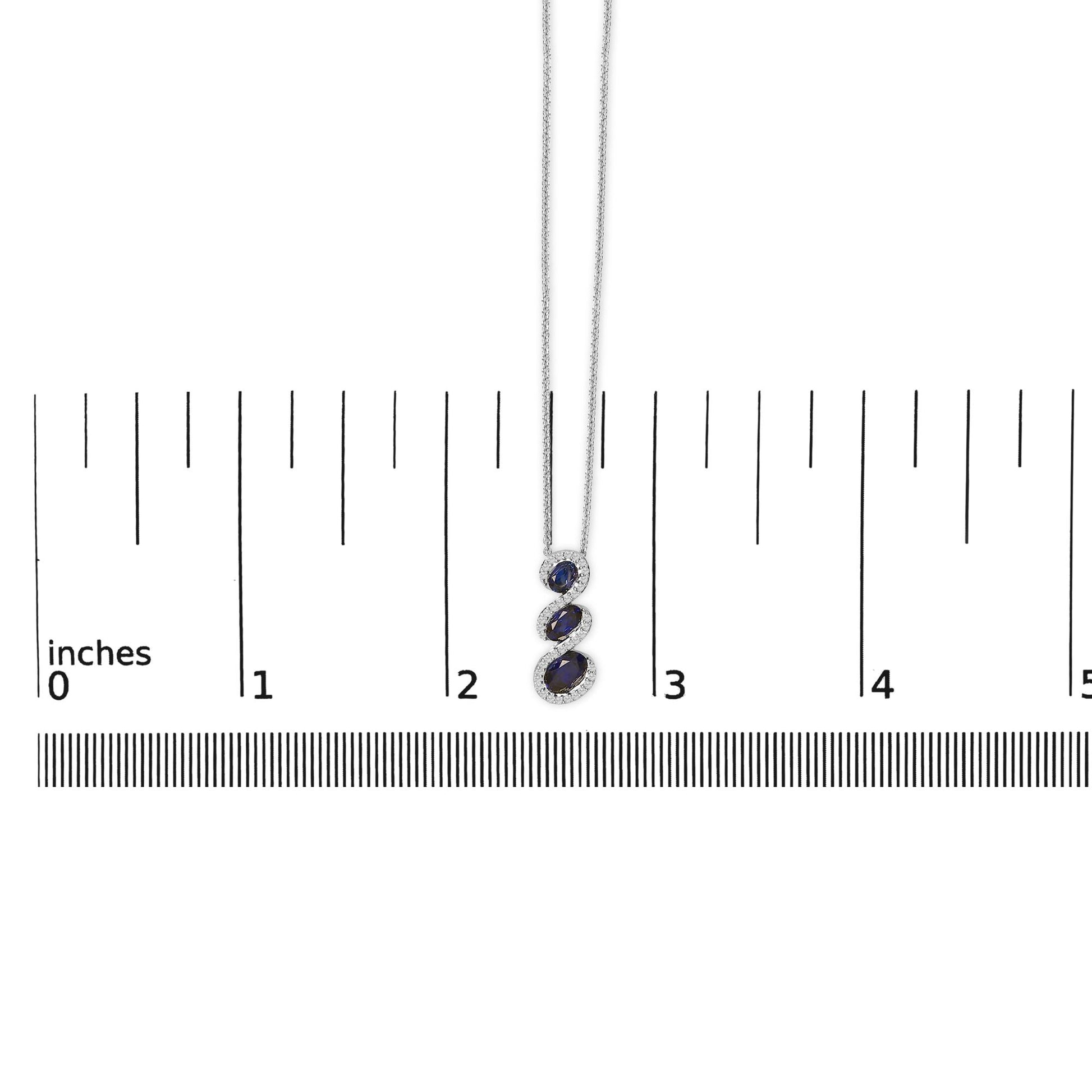 White metal necklace shown on a scale with a vertical pendant featuring three oval blue gemstones surrounded by multiple small round white gemstones in prong settings.