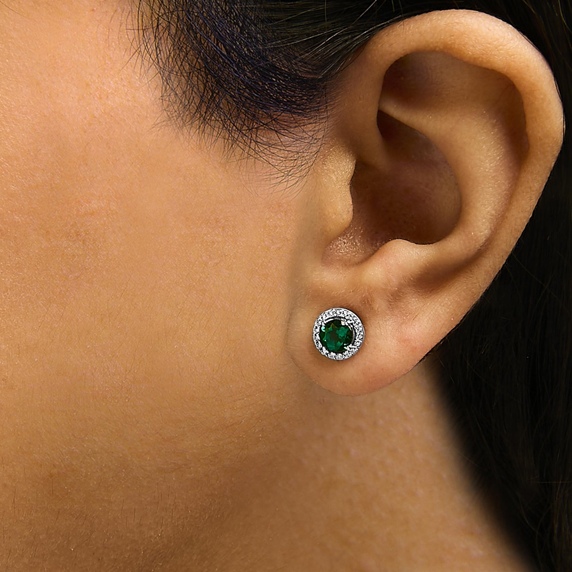 White metal stud earring with a central round green gemstone surrounded by a halo of small white gemstones, shown worn on a model's ear in a close-up side angle.