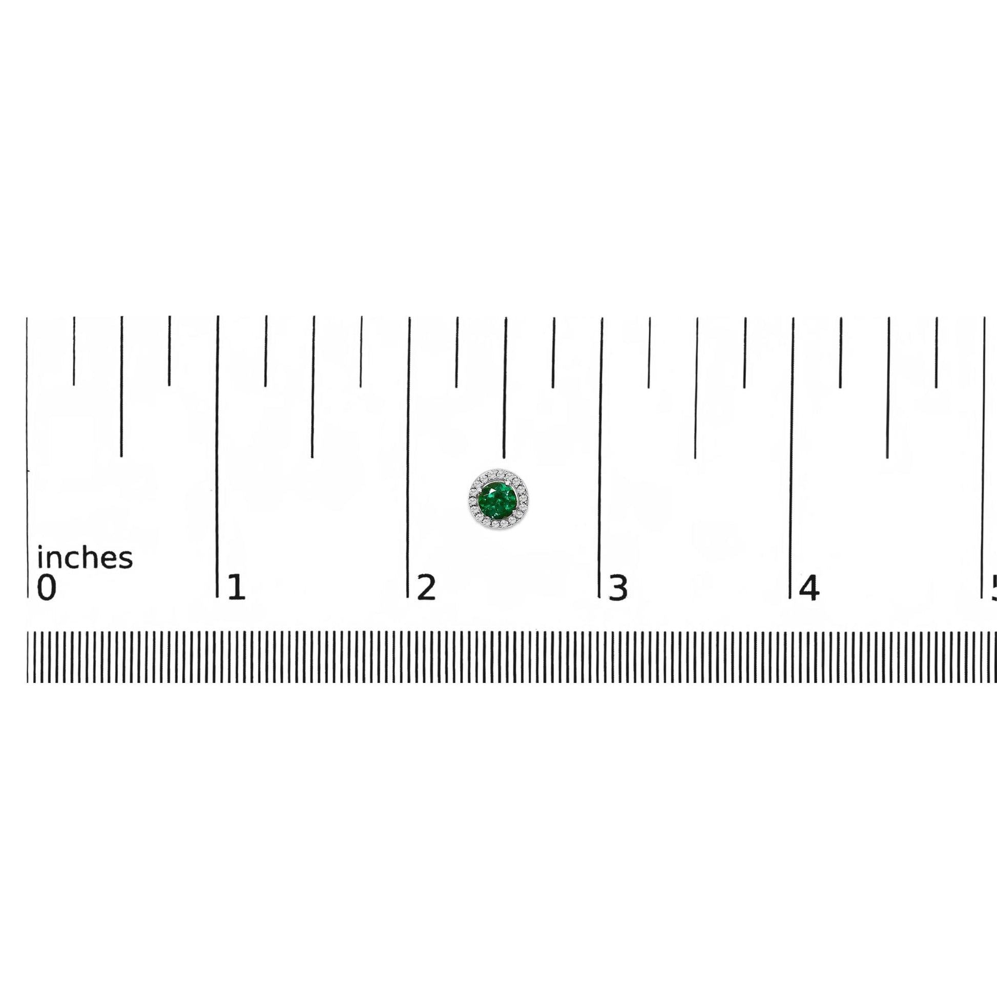 White metal round earring with a green round gemstone center surrounded by small white round gemstones, shown on a ruler scale from a front angle.
