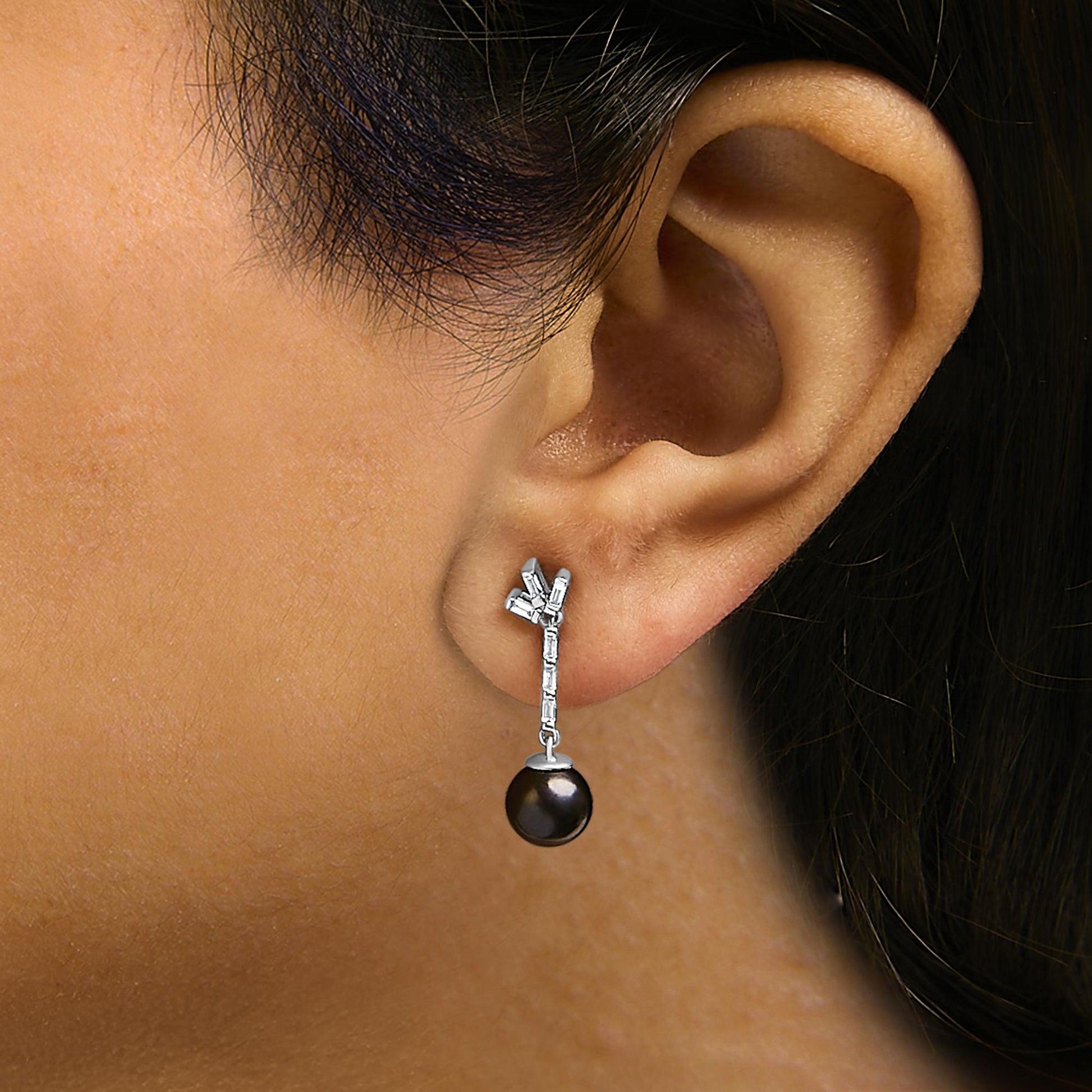 White metal earrings with a vertical arrangement of white baguette-shaped gemstones and a dark round gemstone drop, shown worn on a model in a close-up side view.