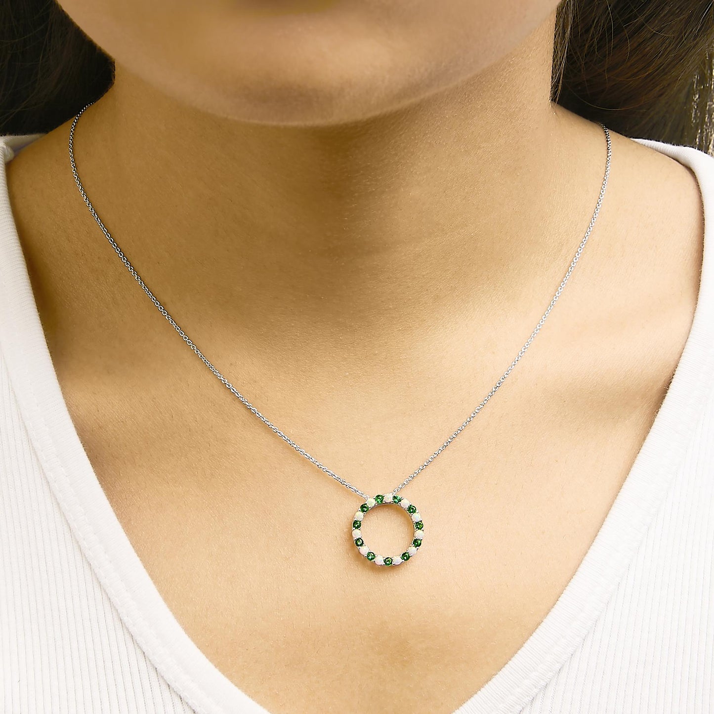 Close-up front view of a white metal necklace worn on a model, featuring a circular pendant set with alternating round green and white gemstones in a shared prong setting.