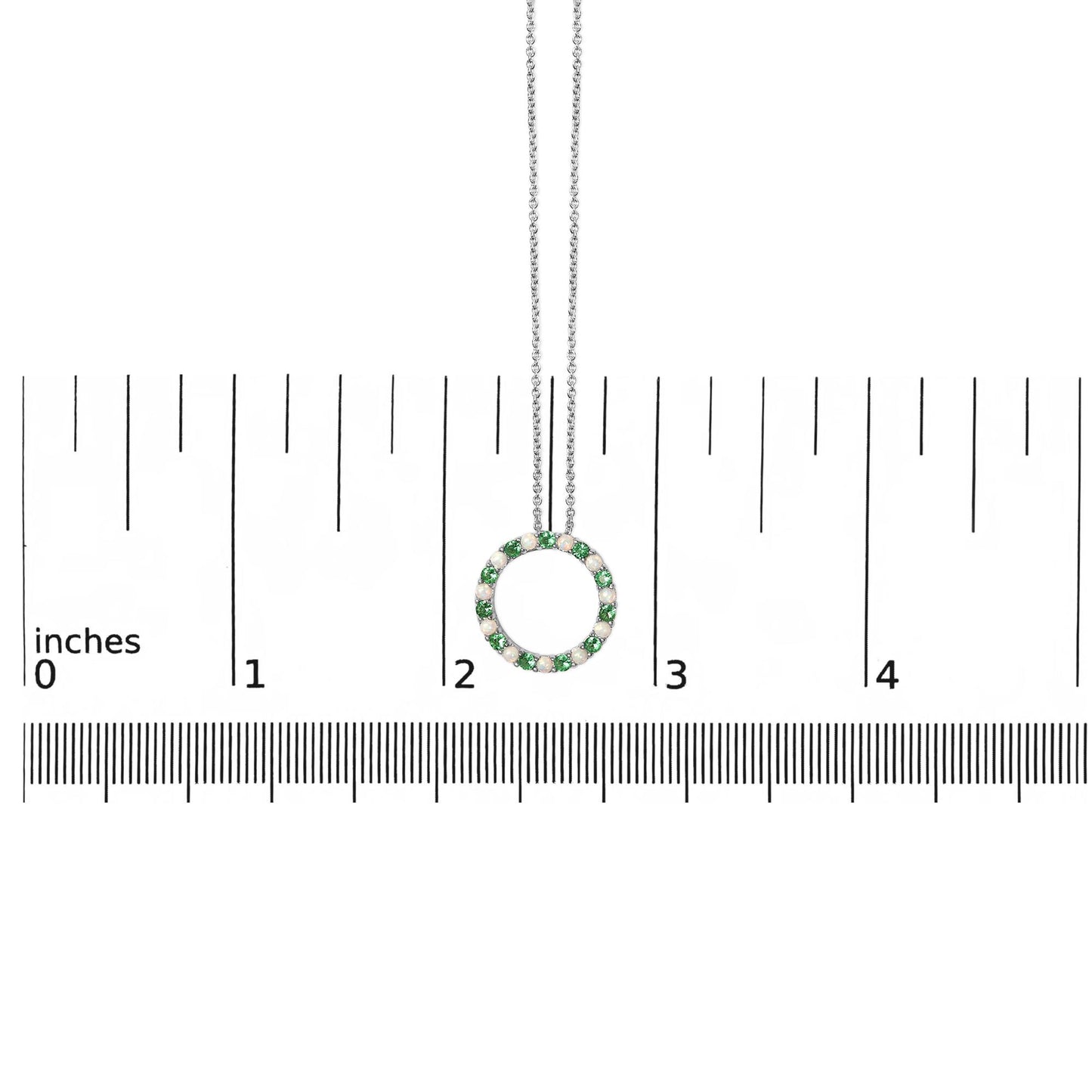 White metal circular necklace pendant with alternating green and white round gemstones in shared prong settings, shown hanging on a chain against a ruler scale from a front angle.