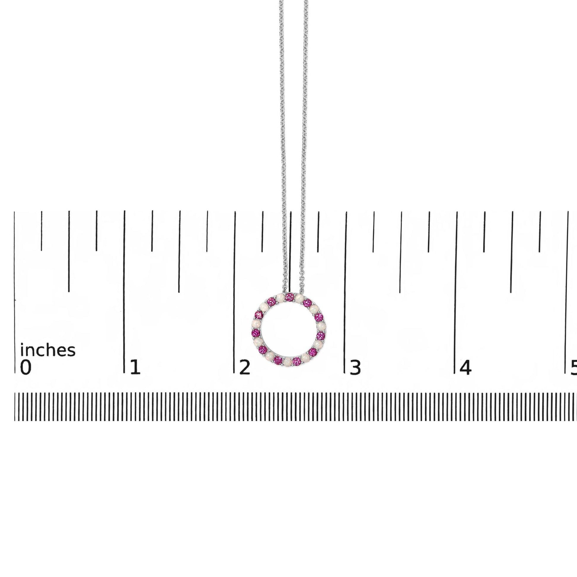 White metal circular necklace pendant featuring alternating round red and white gemstones in prong settings, shown hanging centered on a ruler scale from a front angle.