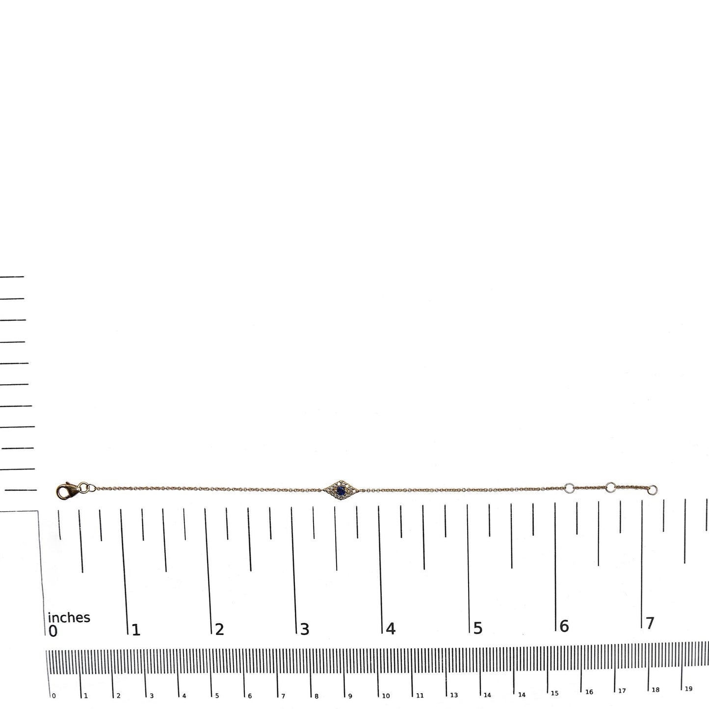 Yellow metal bracelet with a small diamond-shaped centerpiece featuring a blue gemstone surrounded by white gemstones, shown flat on a ruler for size reference, shot from above.