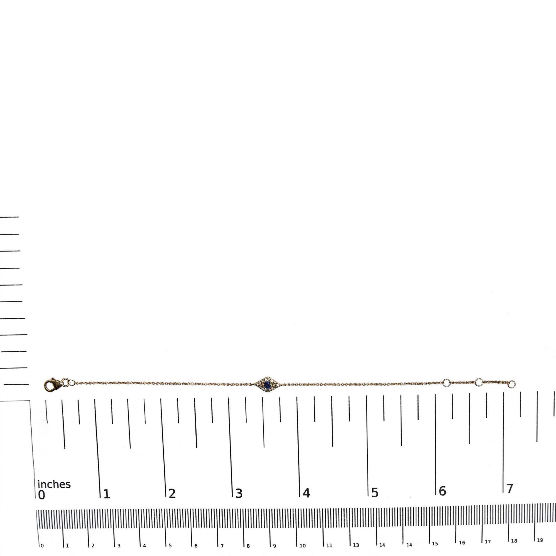 Yellow metal bracelet with a small diamond-shaped centerpiece featuring a blue gemstone surrounded by white gemstones, shown flat on a ruler for size reference, shot from above.