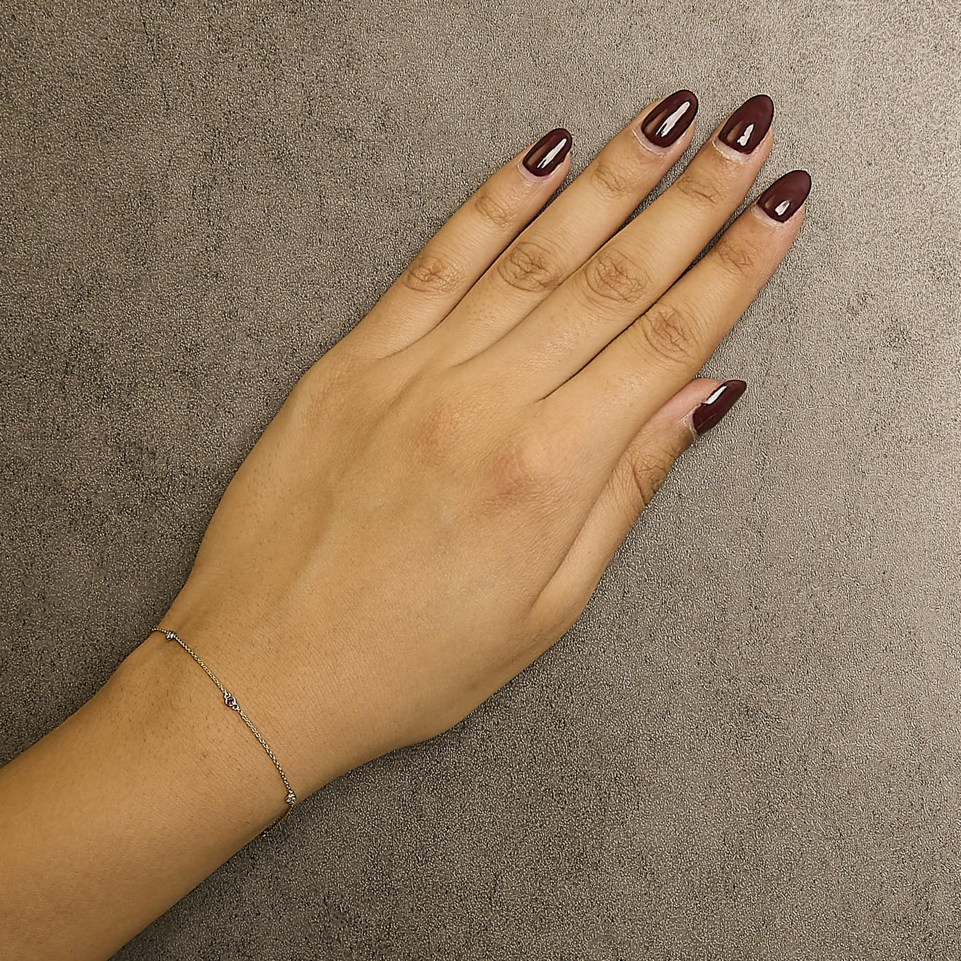 Close-up top view of a yellow metal delicate bracelet with small round red and white gemstones in bezel settings, worn on a model's wrist.