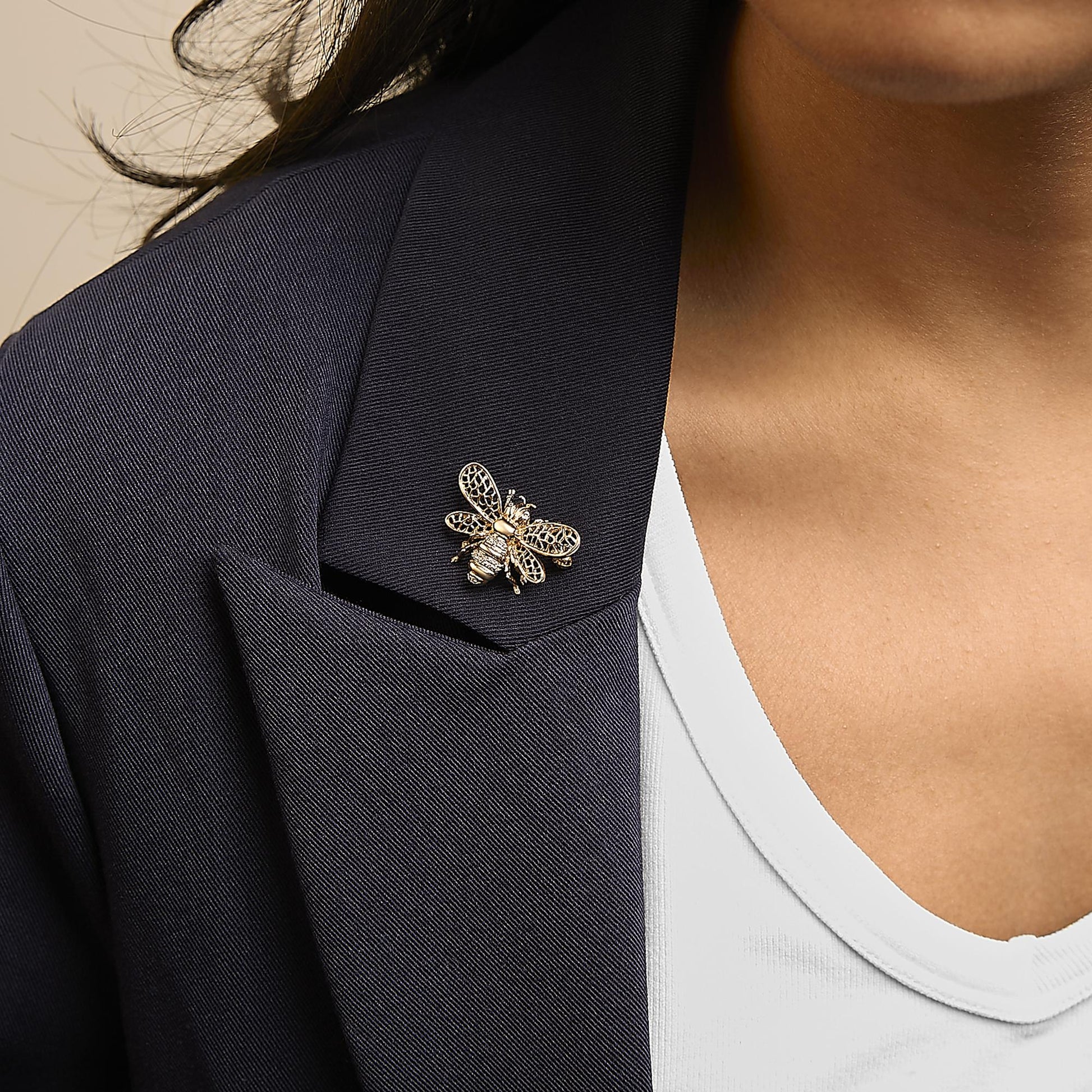 Brooch shaped like a bee with yellow and white metal and white round gemstones in prong settings, shown worn on a model's dark blazer lapel, close-up angled view.