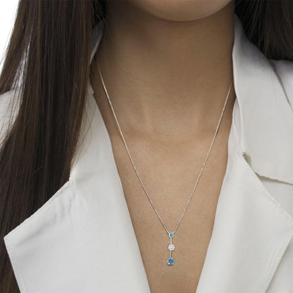 Close-up front view of a white metal necklace on a model, featuring one white gemstone and two blue gemstones in round prong settings arranged vertically.