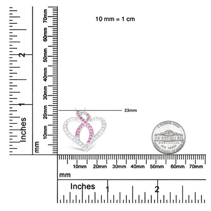 Close-up front view of a white necklace pendant in an open heart shape with intertwined ribbon design, featuring round white and pink gemstones in prong settings, placed on a ruler for size reference.