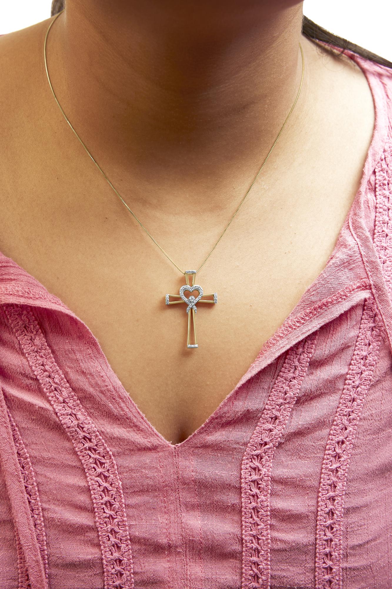 Close-up front view of a yellow metal cross necklace with a heart shape and white round gemstones in prong settings, worn on a model with a pink textured top.
