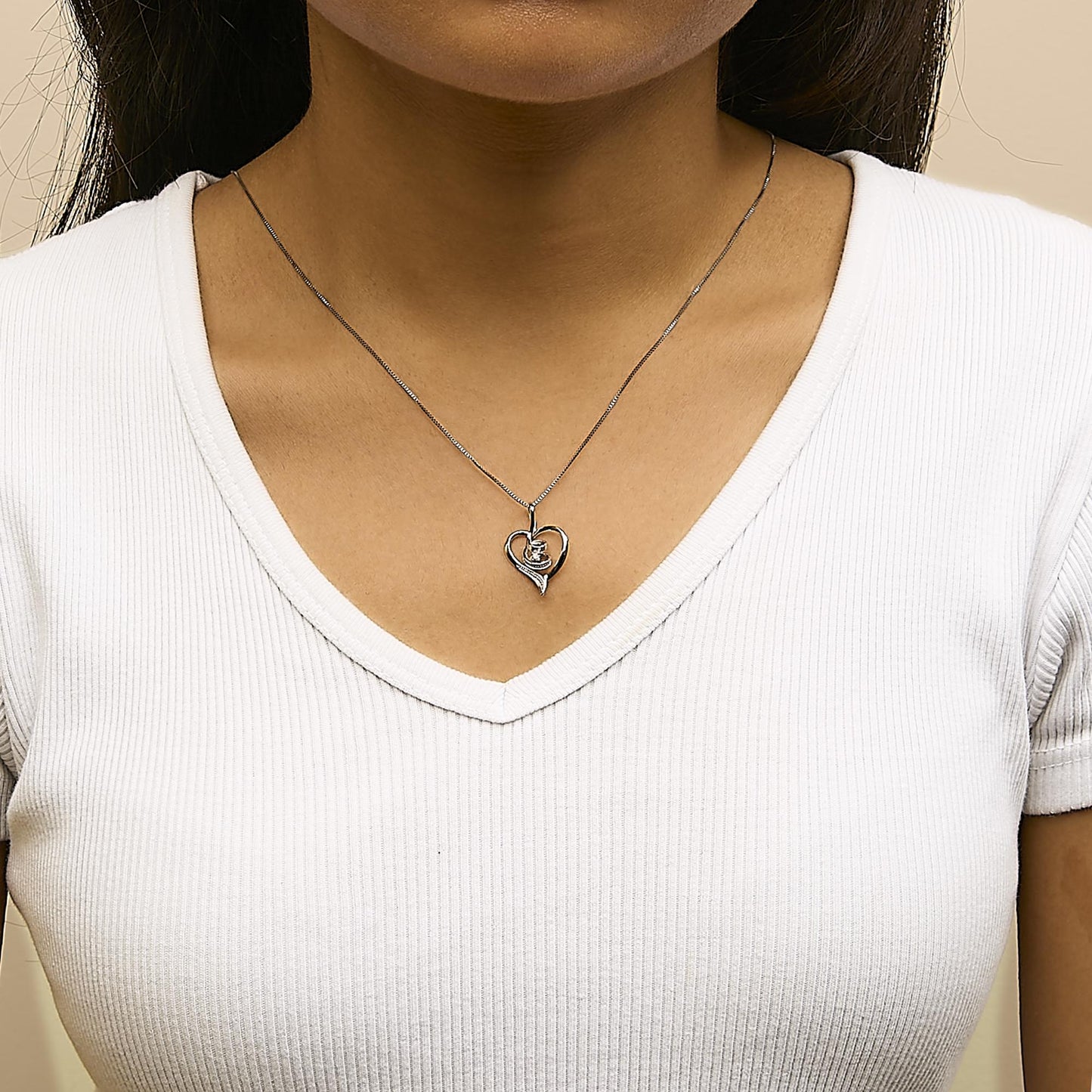 Close-up front view of a white metal heart-shaped necklace pendant with a single round white gemstone in prong setting, worn on a model wearing a white ribbed V-neck shirt.