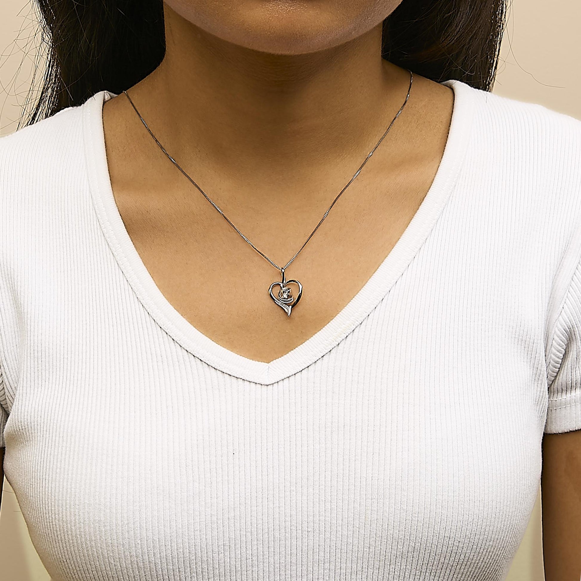 Close-up front view of a white metal heart-shaped necklace pendant with a single round white gemstone in prong setting, worn on a model wearing a white ribbed V-neck shirt.