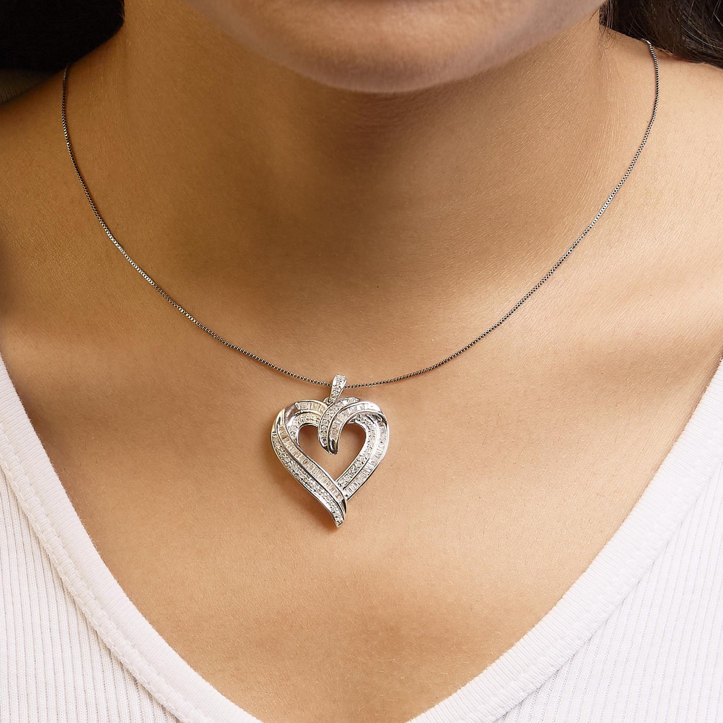 Close-up front view of a white metal heart-shaped necklace pendant with round prong-set white gemstones and baguette channel-set white gemstones, worn on a model.