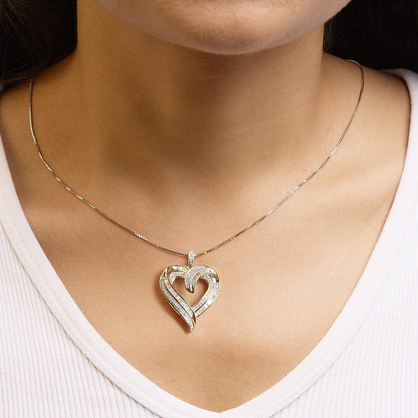 Close-up front view of a yellow metal heart-shaped necklace pendant with white round prong-set gemstones and white baguette channel-set gemstones, worn on a model.