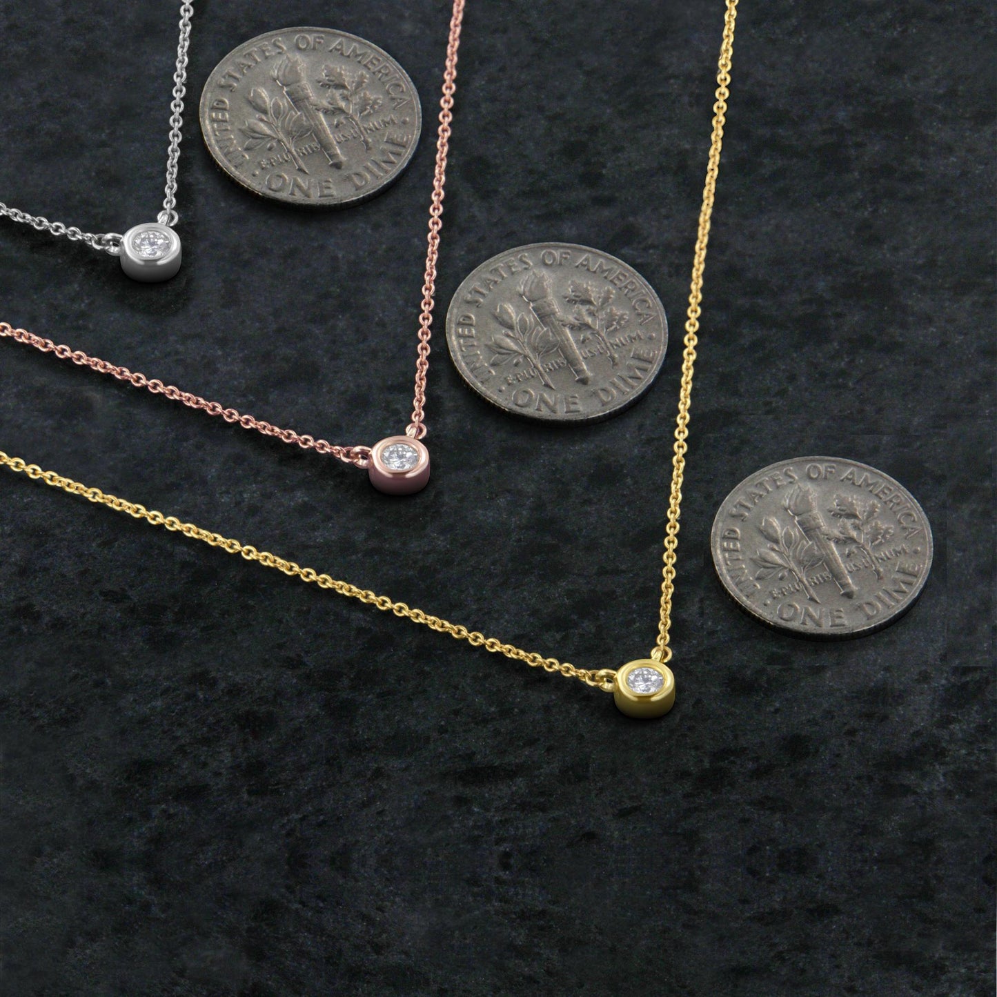 Close-up top view of three necklaces in yellow, rose, and white metal, each featuring a single round white gemstone in a bezel setting, displayed next to coins for size reference.