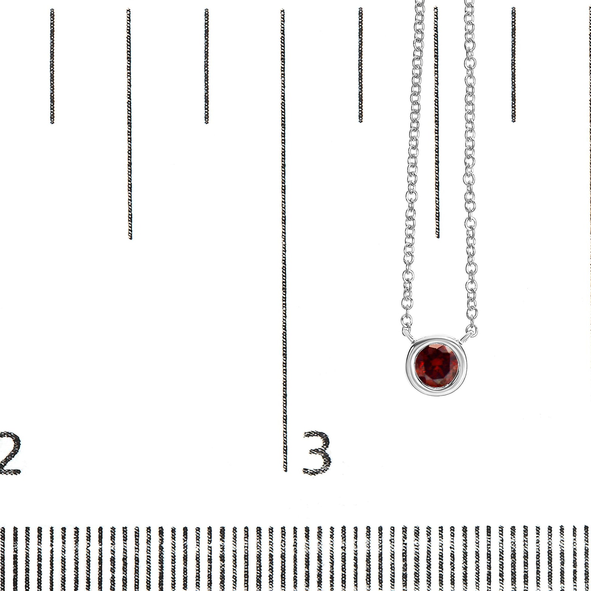 Close-up front view of a white metal necklace with a round red gemstone in a bezel setting, shown on a ruler for size reference.