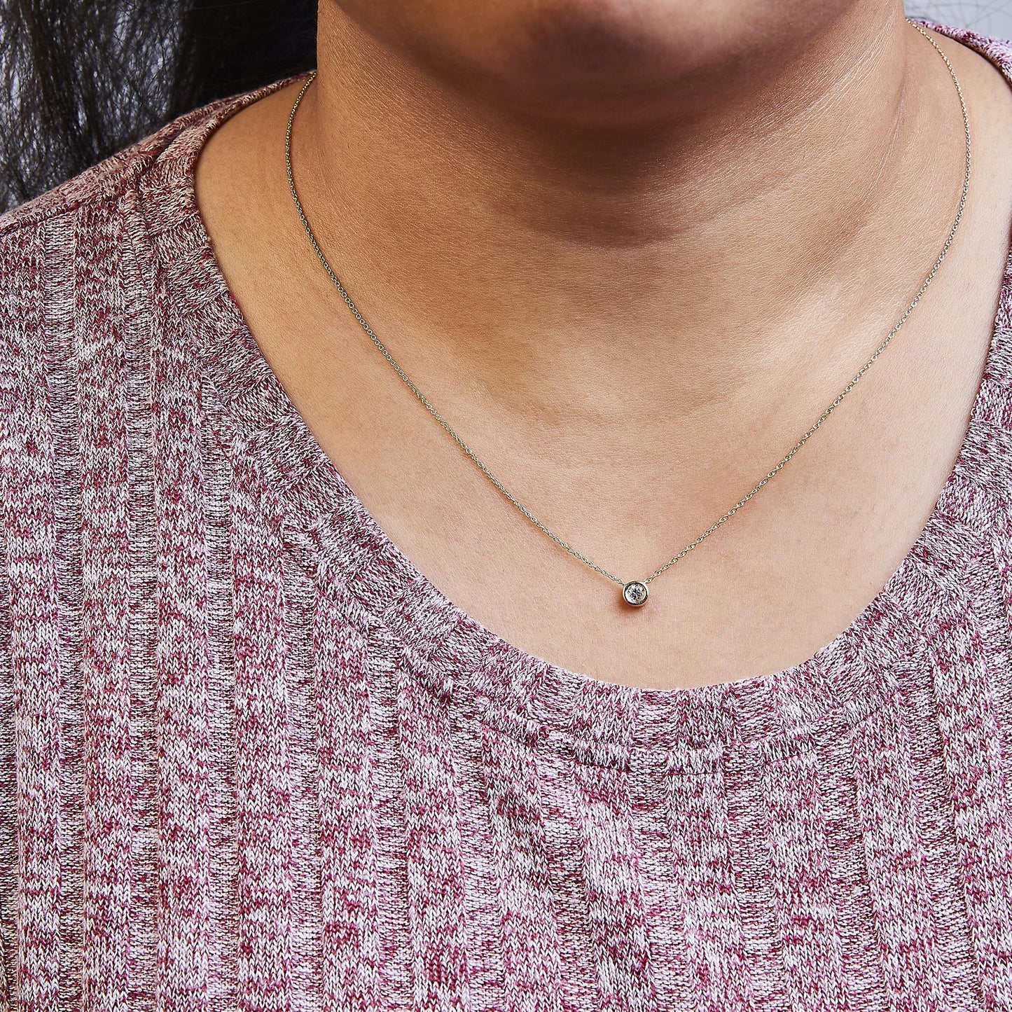 Close-up front view of a yellow metal necklace with a single round white gemstone in a bezel setting, worn on a model with a pink textured top.