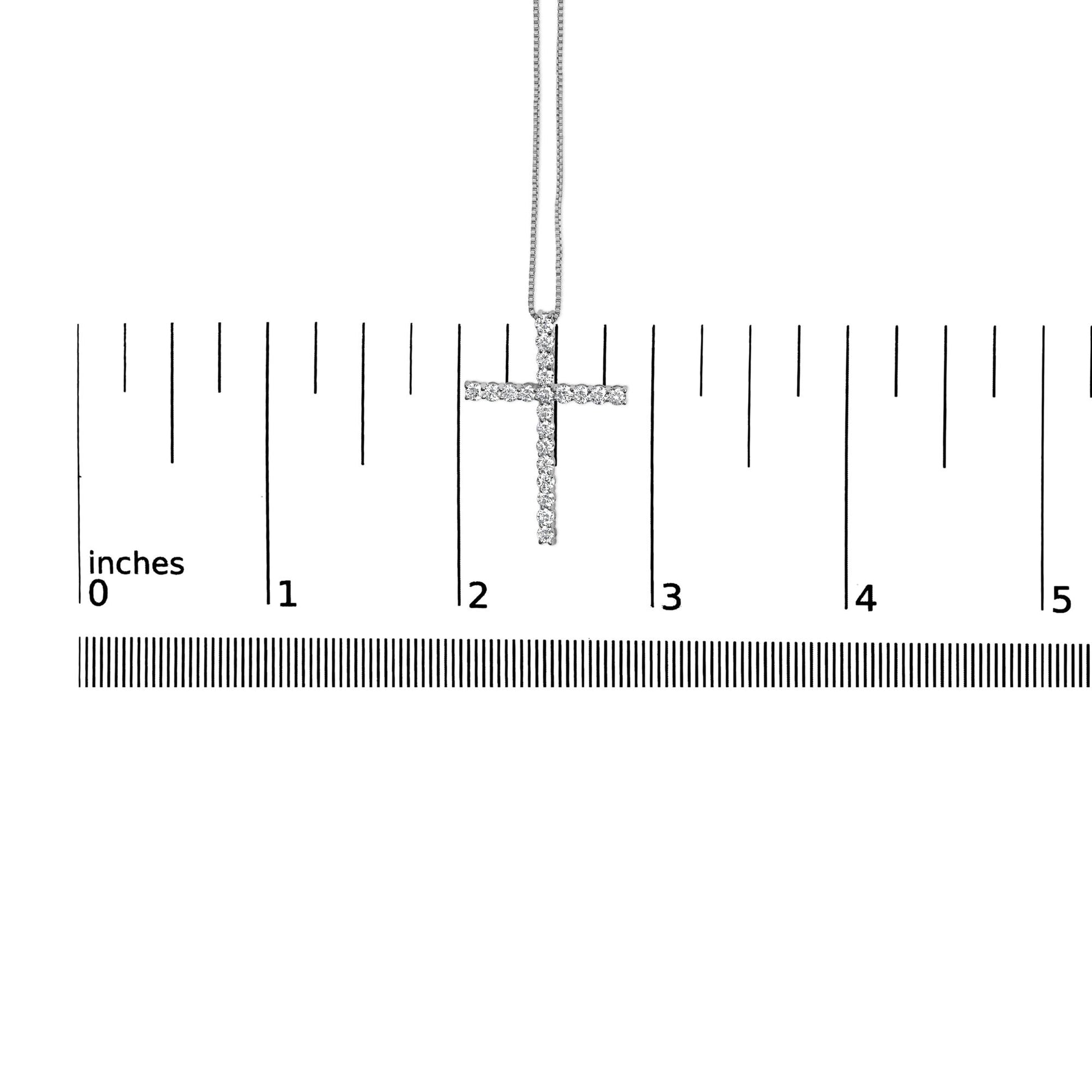 White metal cross necklace with round white gemstones in prong setting, hanging vertically on a ruler for size reference, front view.