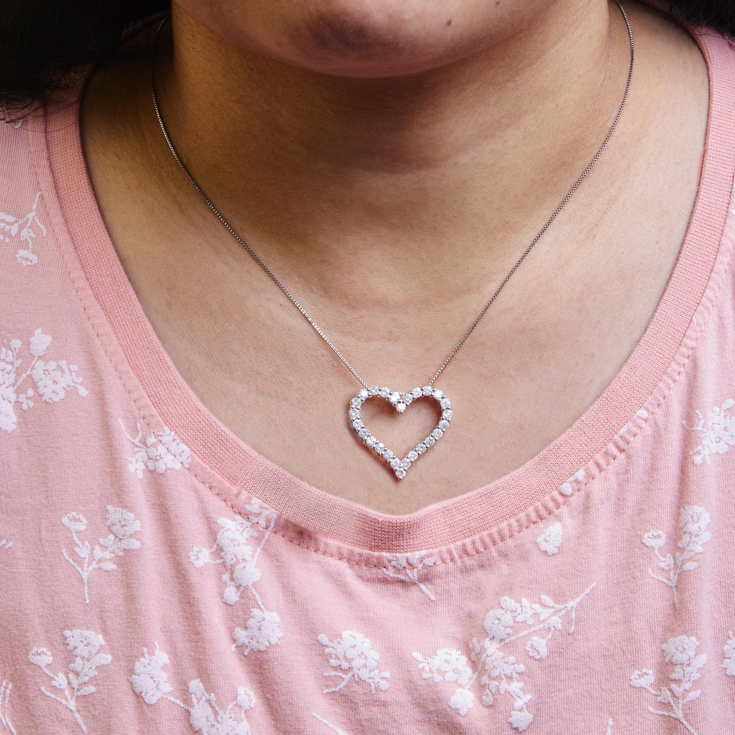 Close-up front view of a necklace on a model with a heart-shaped pendant featuring round white gemstones set in shared prongs on a white metal chain.