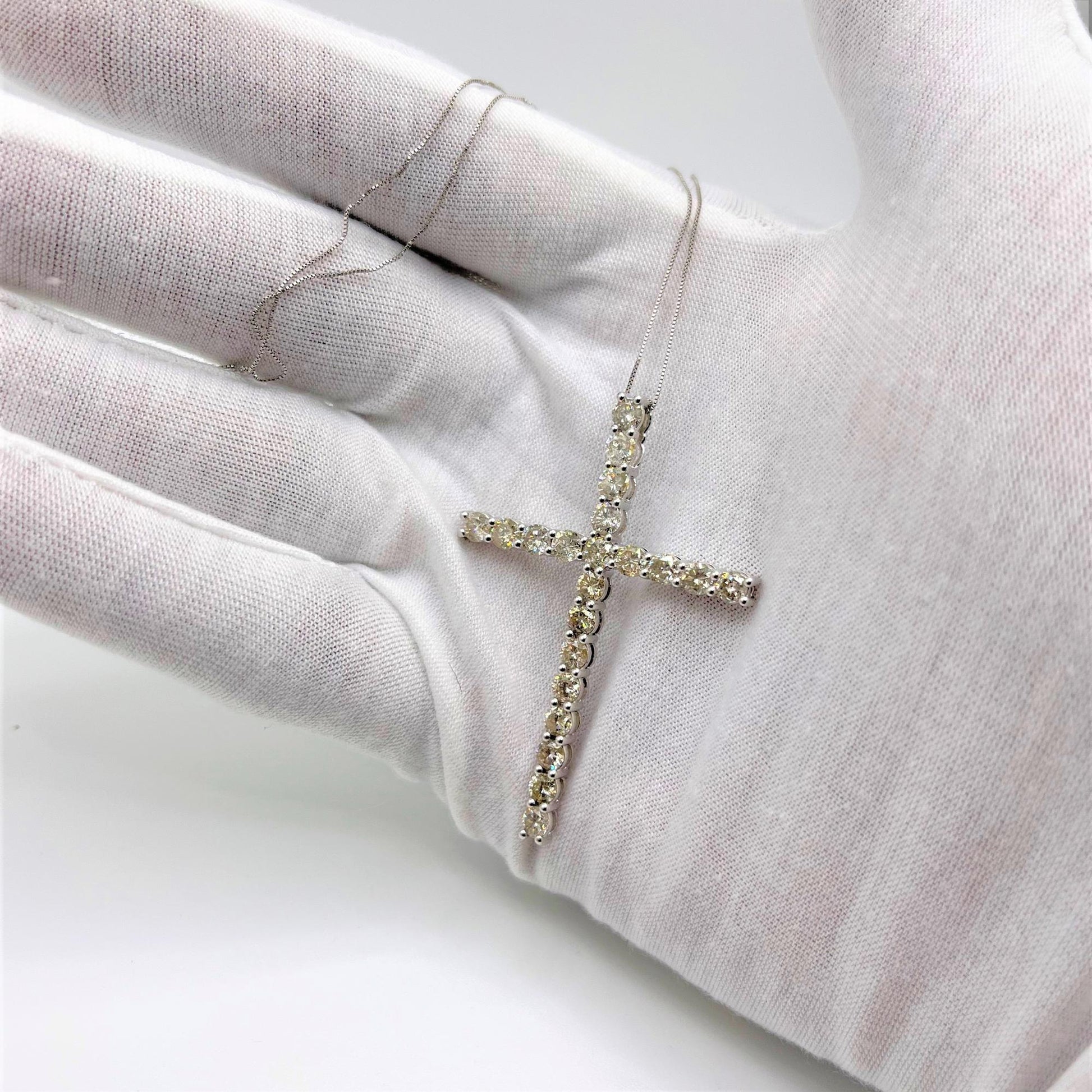 Close-up angled view of a white metal cross necklace with round white gemstones in prong settings, shown being held on a white-gloved hand.